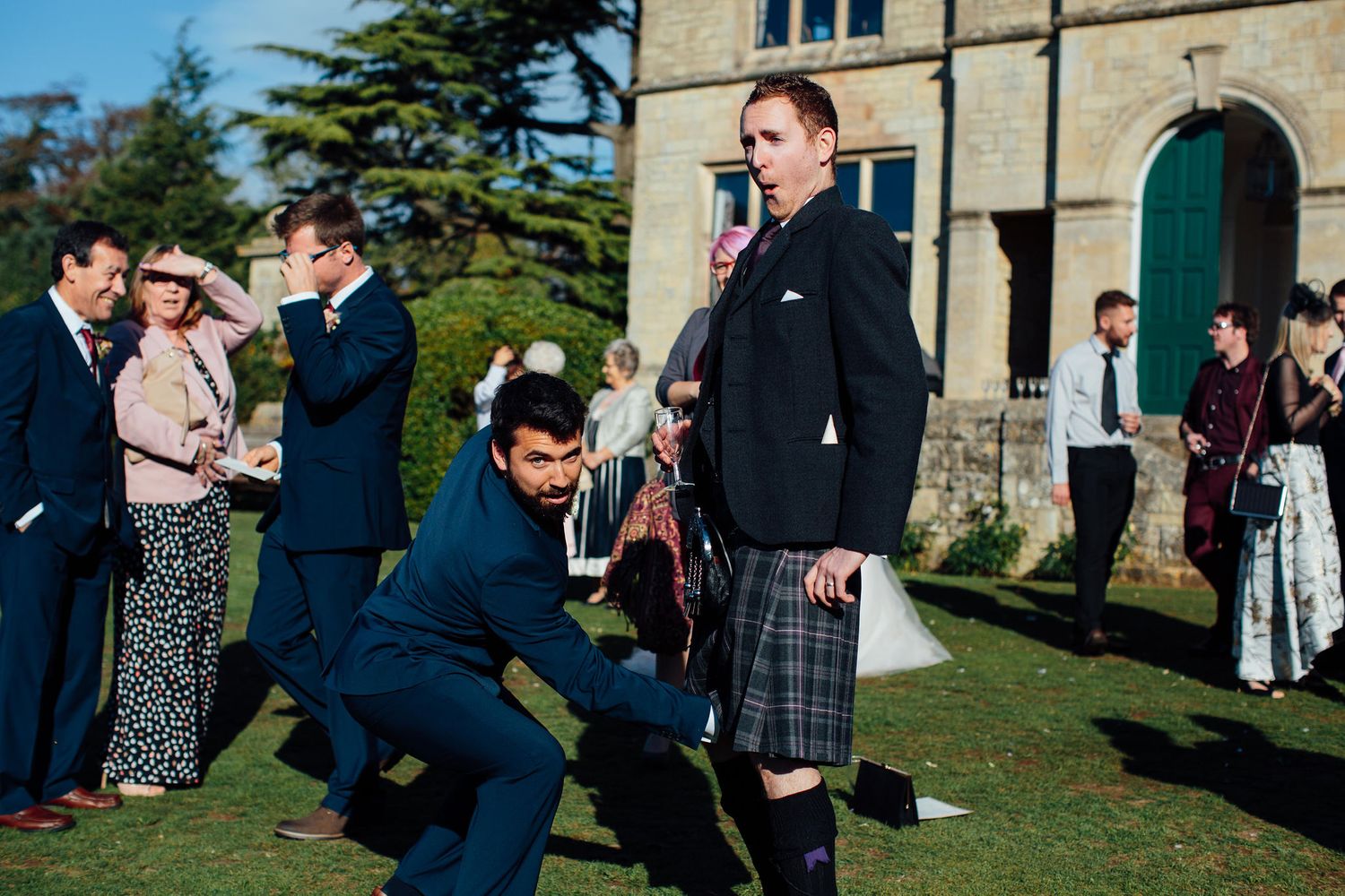 Wedding guests mingle on a sunny lawn outside a grand stone building.