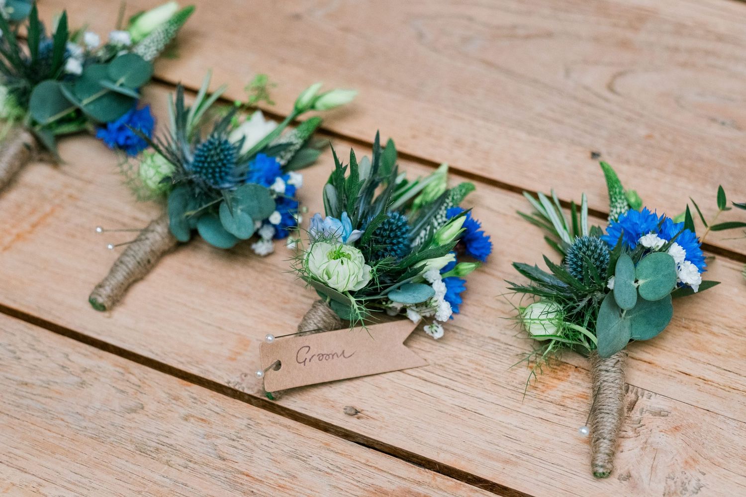 Blue and green wedding boutonnieres with twine wrapping laid out on wooden surface.