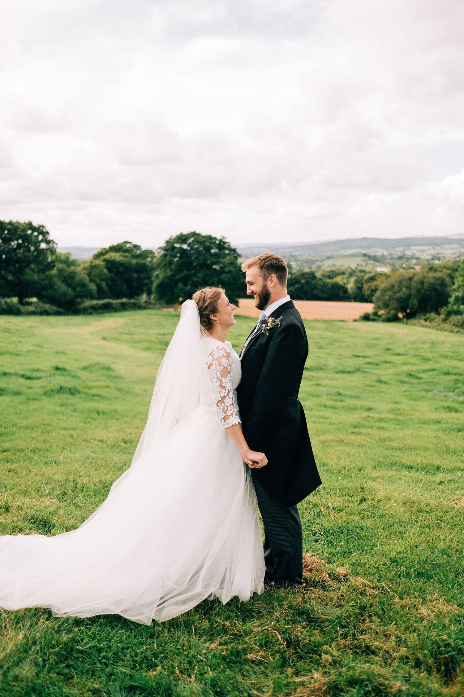 Newlyweds walking through a scenic green field with rolling hills in the background.