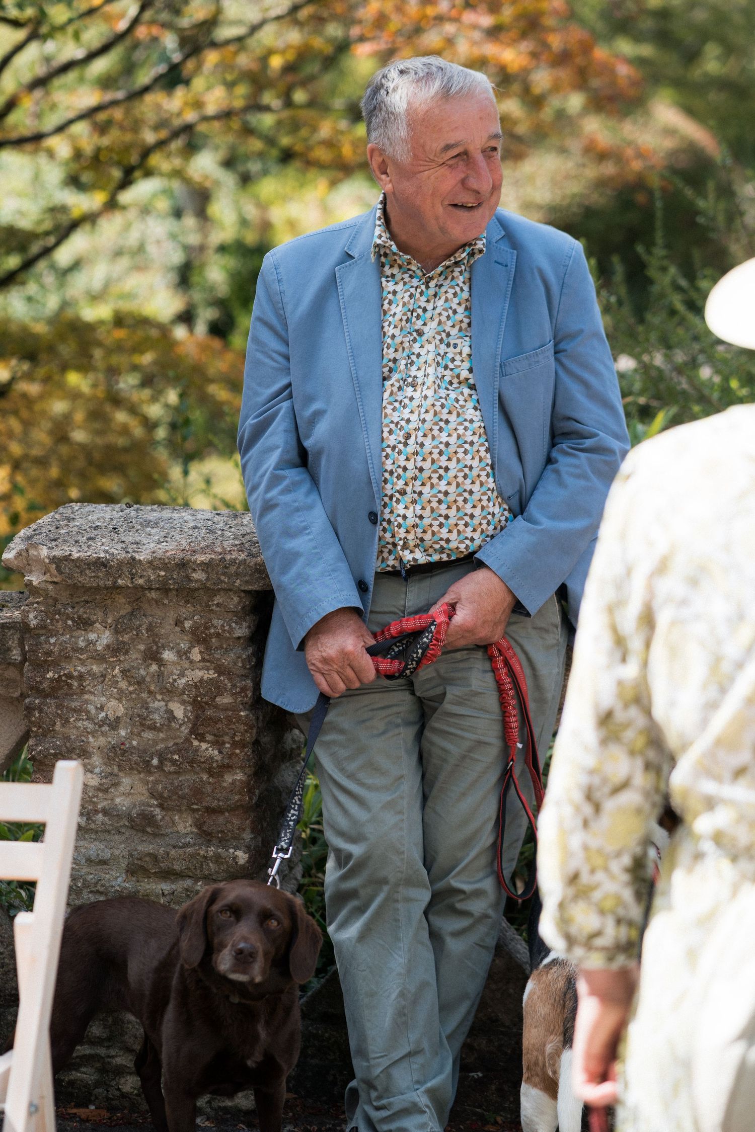 Smiling older man in blue blazer holding red dog leash with chocolate Labrador at outdoor garden event.