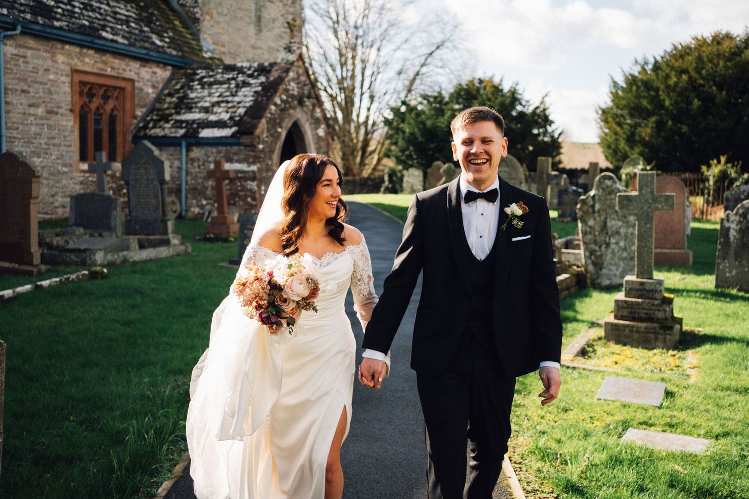 Bride and groom holding hands walking through a church graveyard on their wedding day in natural sunlight.