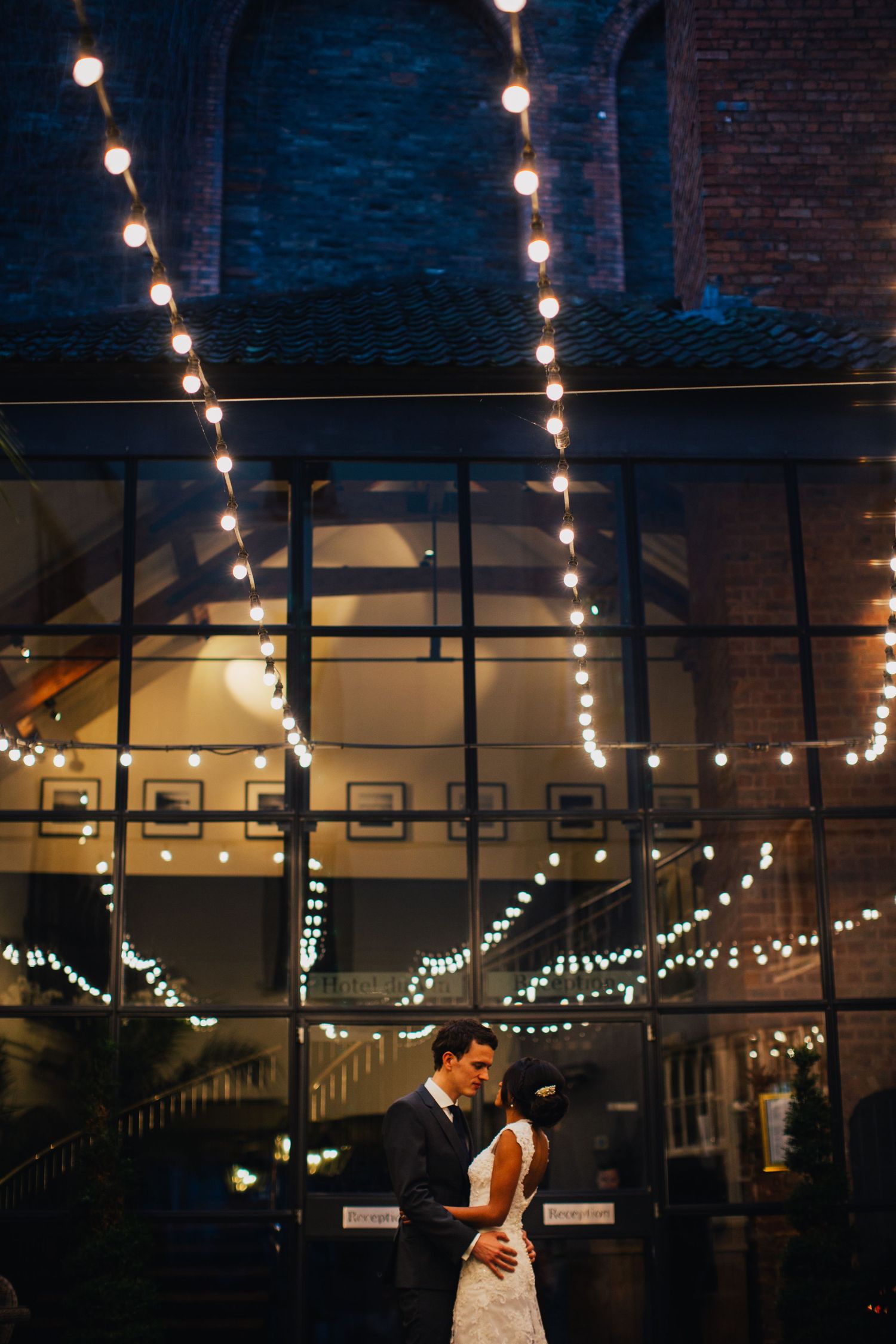 A romantic evening wedding scene with string lights illuminating a dark brick venue as a couple shares an intimate moment.