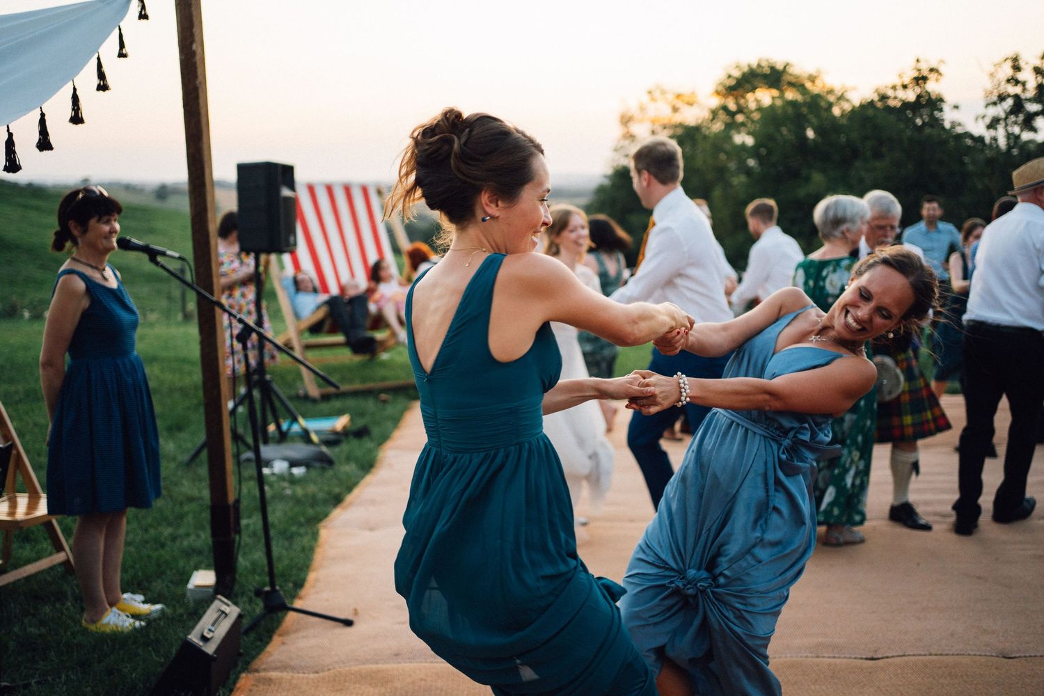 Wedding guests in matching teal dresses dance joyfully on an outdoor patio at sunset.