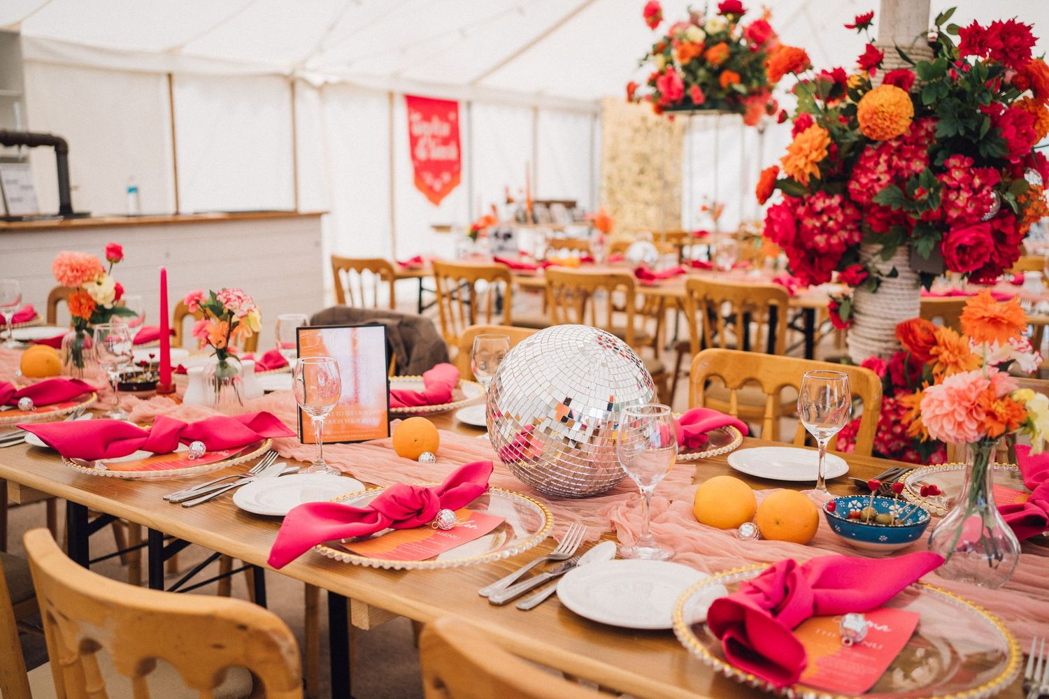 Wedding reception table decorated with vibrant pink and orange floral arrangements and place settings in a white tent.