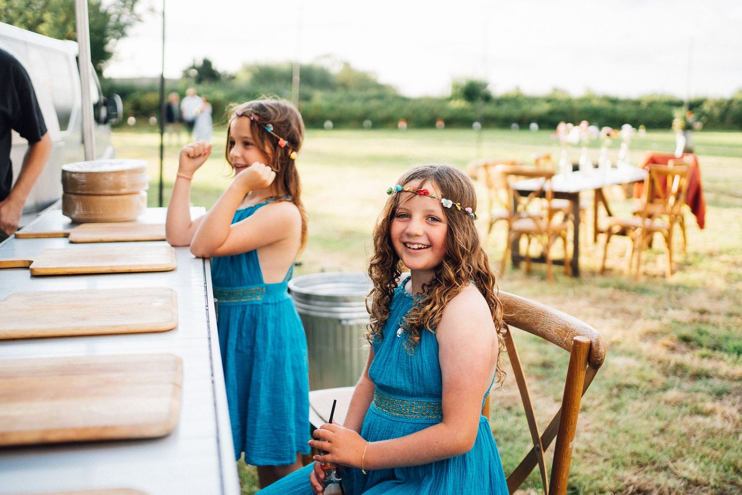 A wedding reception takes place outdoors with rustic wooden tables and chairs set up in a field at sunset.
