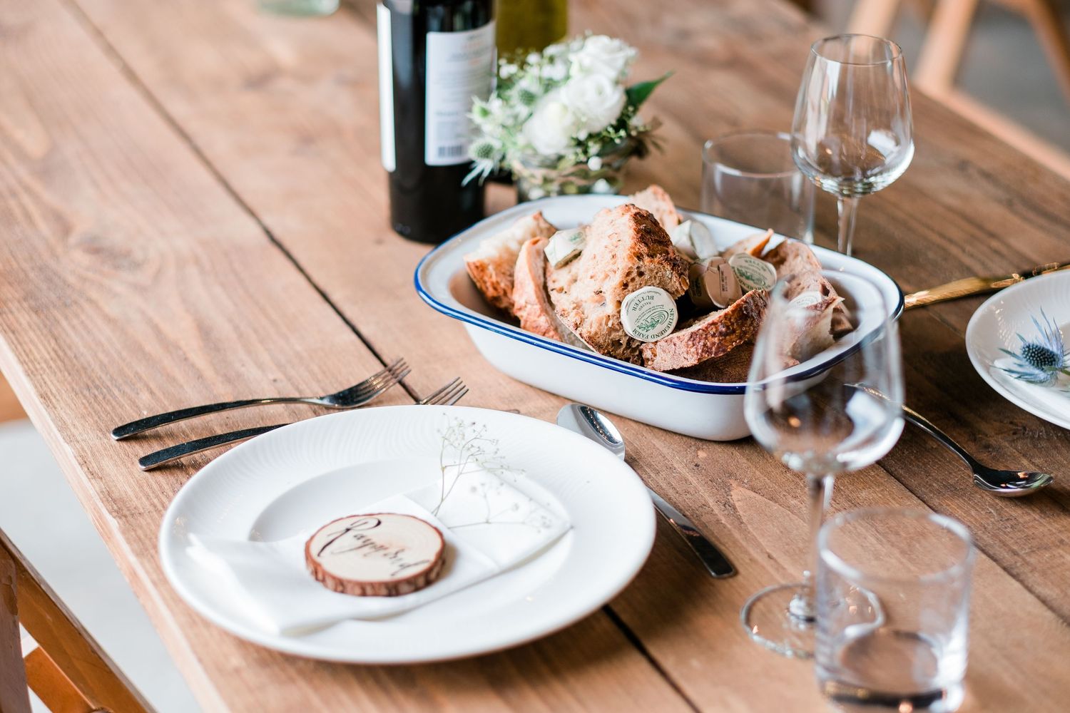 Elegant place settings with wine glasses and plates on wooden tables at a rustic reception.