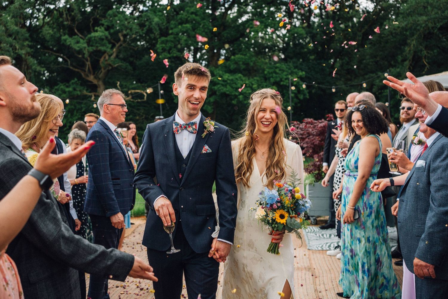 Wedding guests celebrating and tossing confetti over newlyweds in an outdoor garden setting.