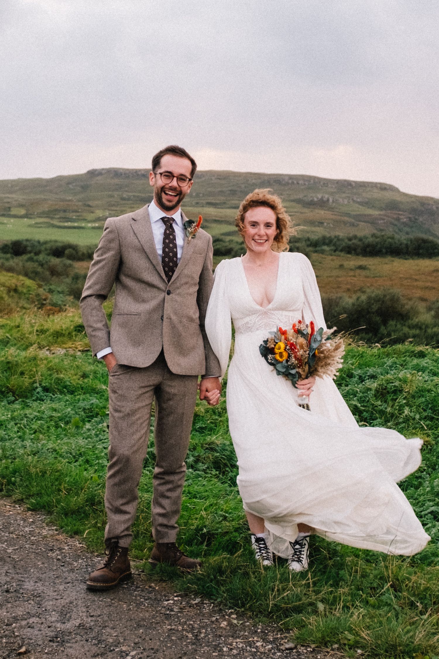 A smiling bride and groom hold hands on a rural path, she in a white gown with sneakers, he in a tan suit.