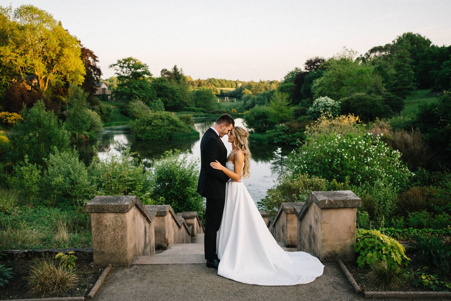 Bride and groom share a romantic kiss on stone steps overlooking a serene garden lake at golden hour.
