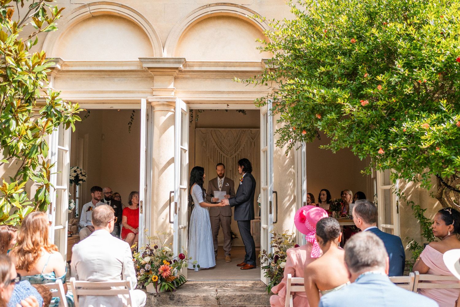 Outdoor wedding ceremony at elegant venue with couple exchanging vows, guests seated, floral arrangements, and lush greenery.