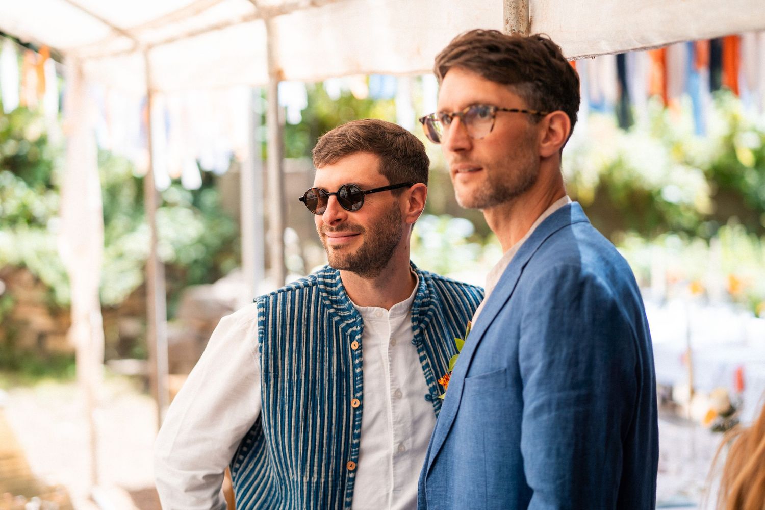 Two stylish men in summer outfits smile at an outdoor festival, one in sunglasses and striped vest.