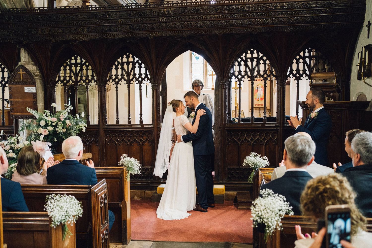 Intimate church wedding ceremony with guests seated in wooden pews and decorative floral arrangements.