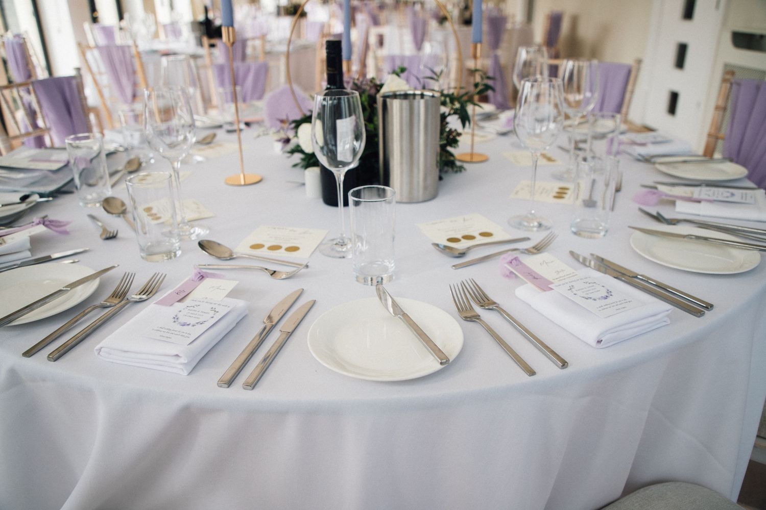 Formal table setting with white linens, silverware, and purple accents around a wine chiller centerpiece.