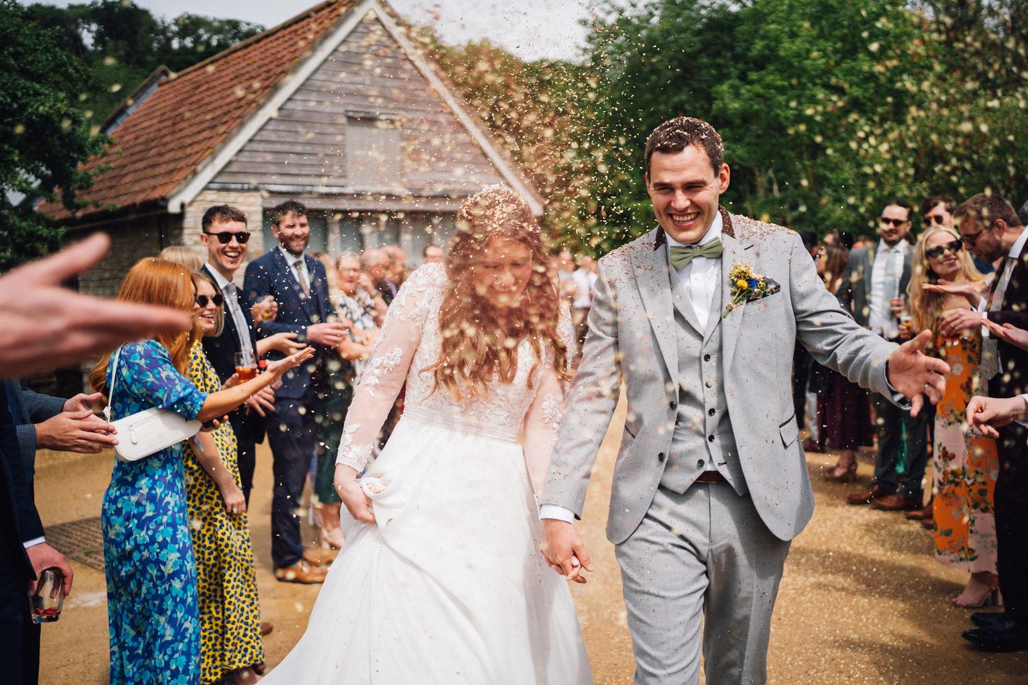 Wedding guests shower confetti on newlyweds walking down path outside rustic barn venue.