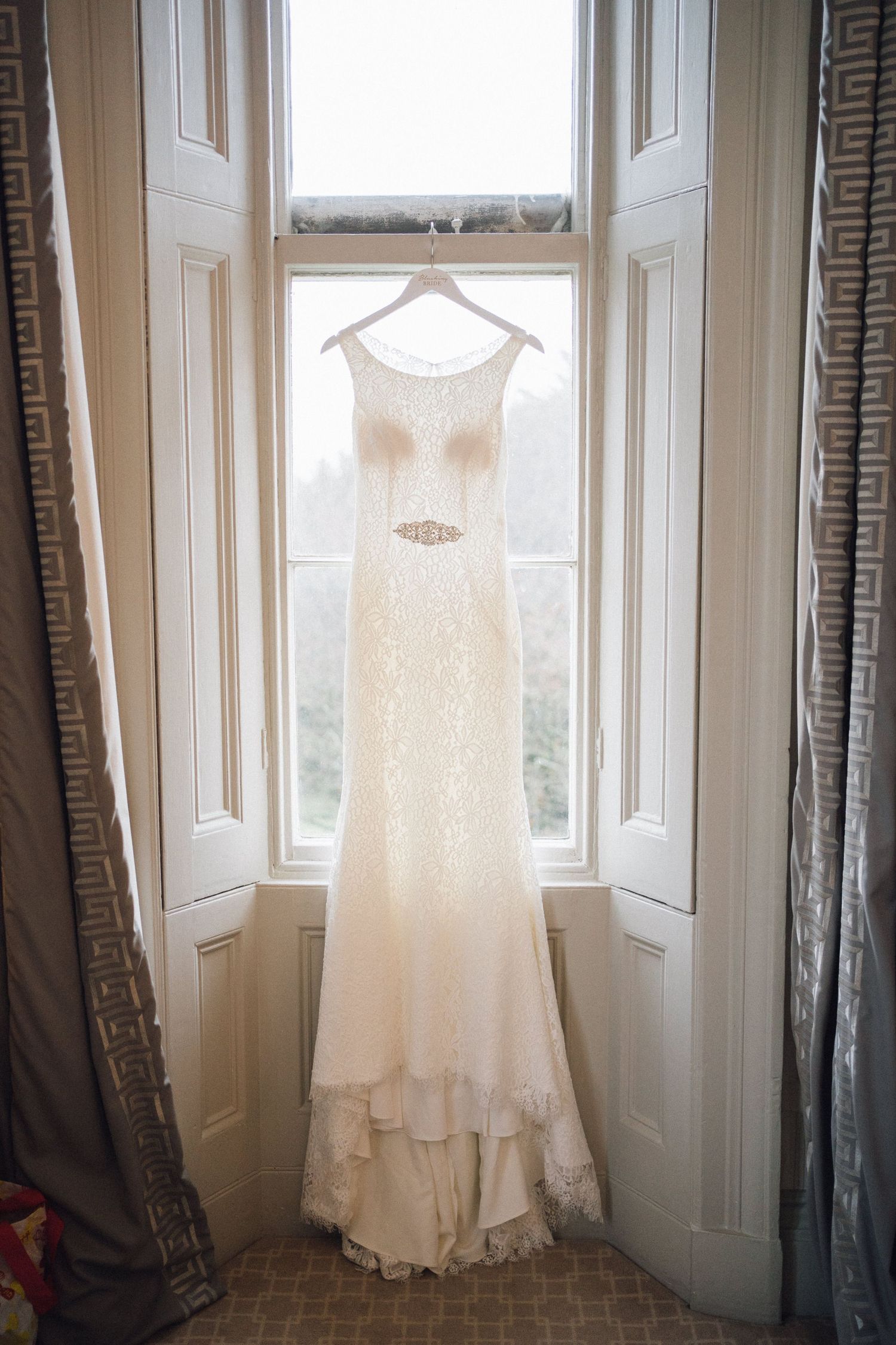 White wedding dress hanging in front of bright bay windows with natural light streaming in.