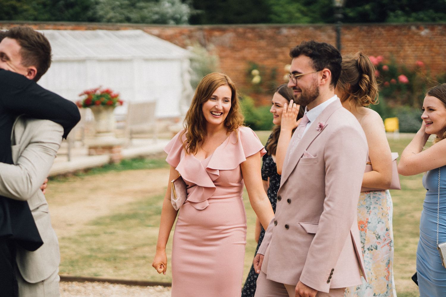 Wedding guests mingle and socialize during an outdoor garden reception near a brick wall.