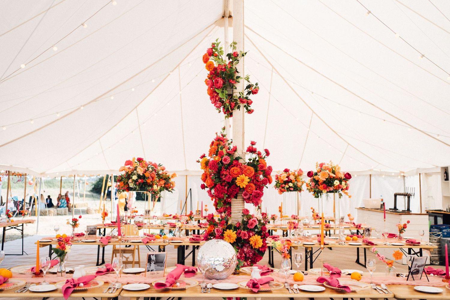 Elegant wedding reception setup inside white tent with pink and coral floral centerpieces and chandeliers overhead.