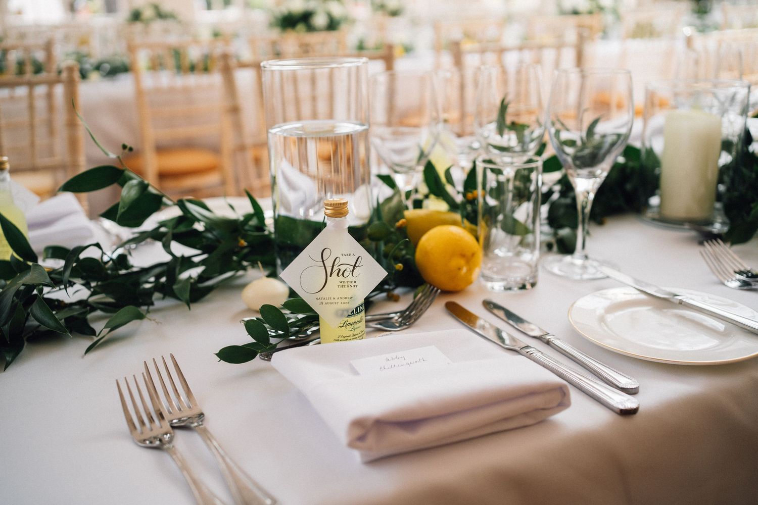 Elegant wedding reception table settings with white linens, glassware, and greenery in a bright indoor venue.
