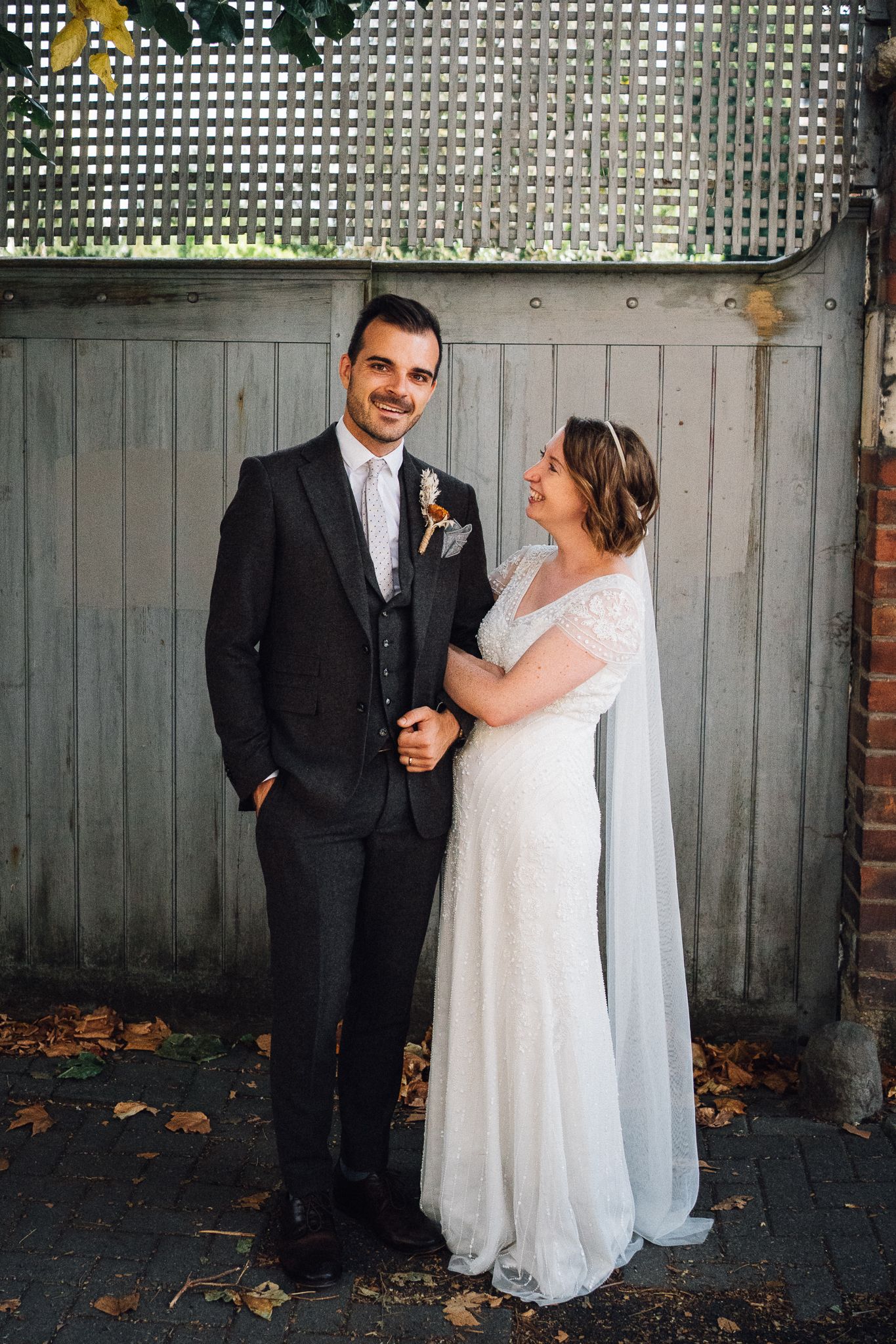 A bride and groom share a candid moment together against a grey fence in an outdoor garden setting.