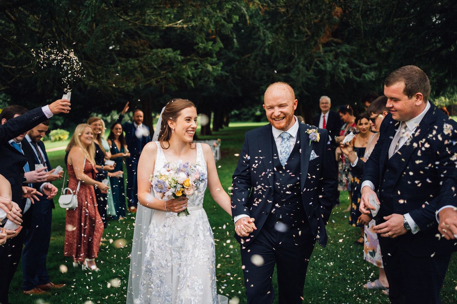 Bride and groom walk through confetti shower surrounded by smiling wedding guests in a lush garden setting.