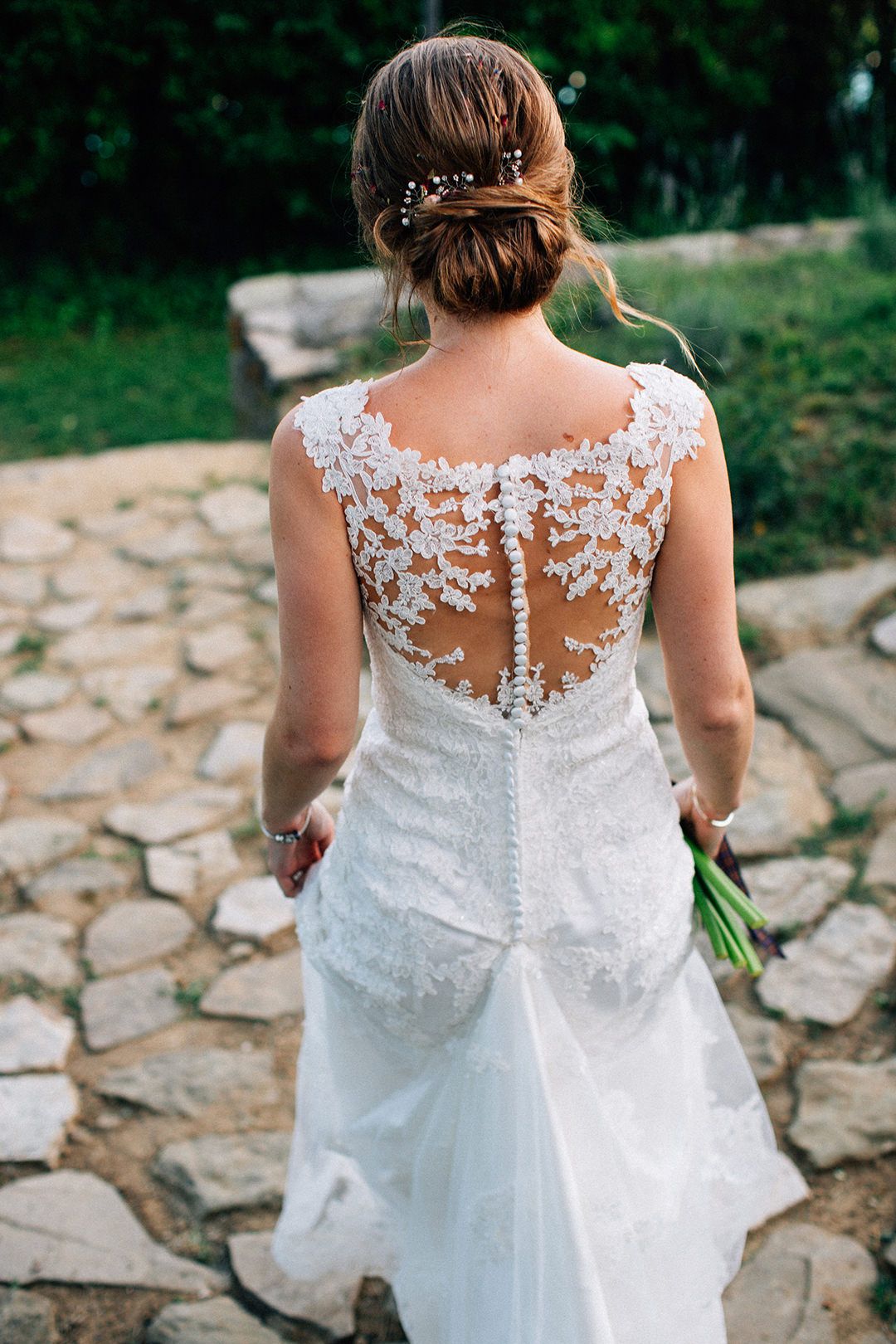 Back view of a bride in a white lace wedding dress with sheer illusion details walking on stone path outdoors.