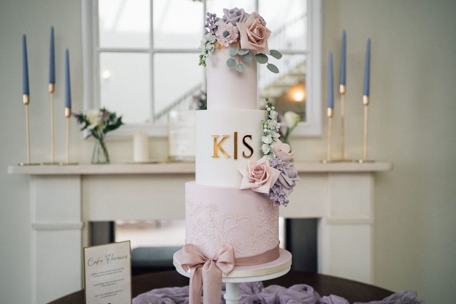 Elegant blush pink wedding cake decorated with delicate sugar flowers and metallic gold accents on white display stand.