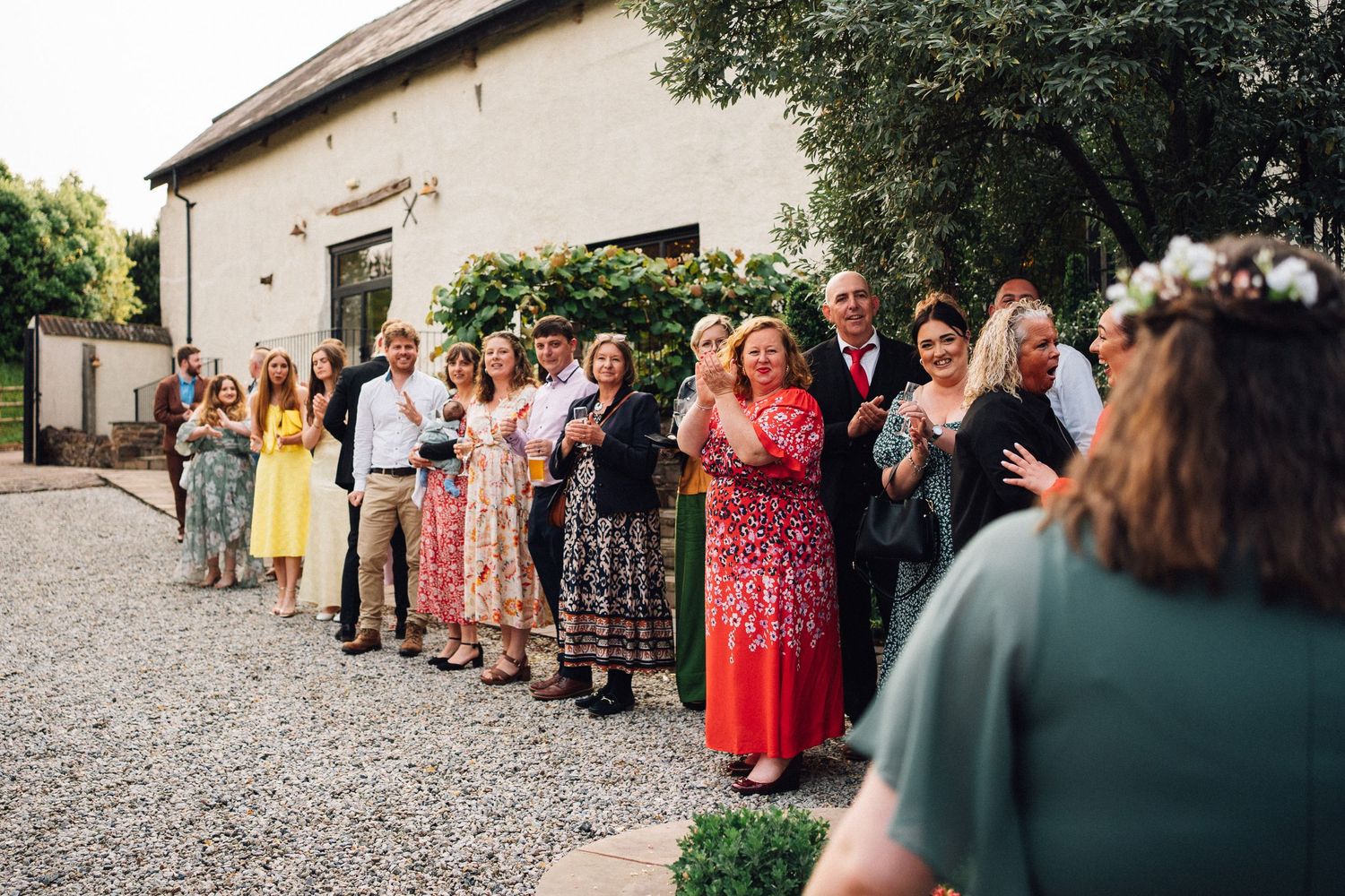 A line of wedding guests in colorful dresses and formal attire stand outside against a stone building wall.