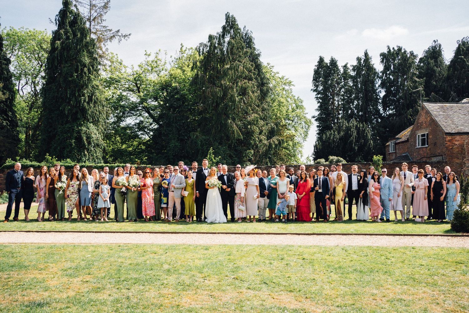 Large group wedding photo of guests lined up on a sunny lawn with evergreen trees in background.
