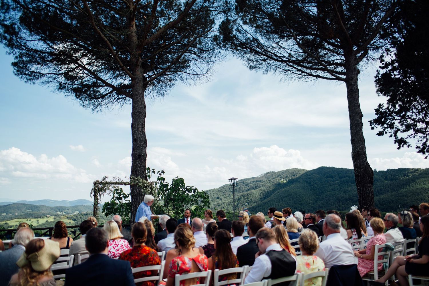 Wedding guests seated outdoors under pine trees with mountain landscape views.