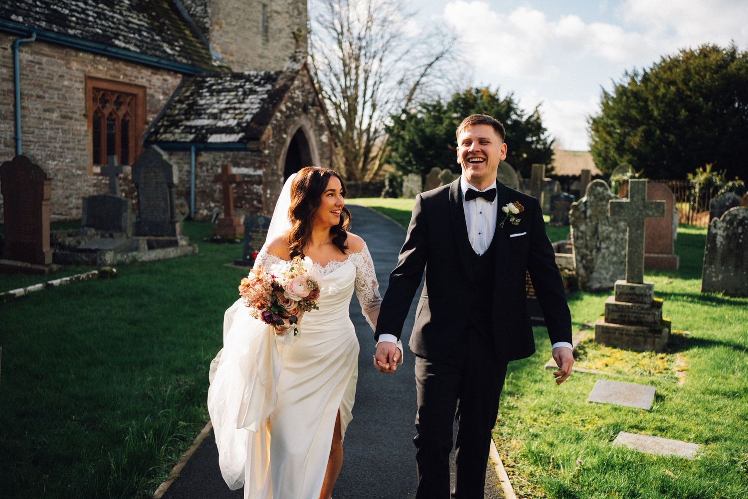Newlywed couple walking hand in hand outside a stone church, bride in white gown with bouquet, groom in black tuxedo.