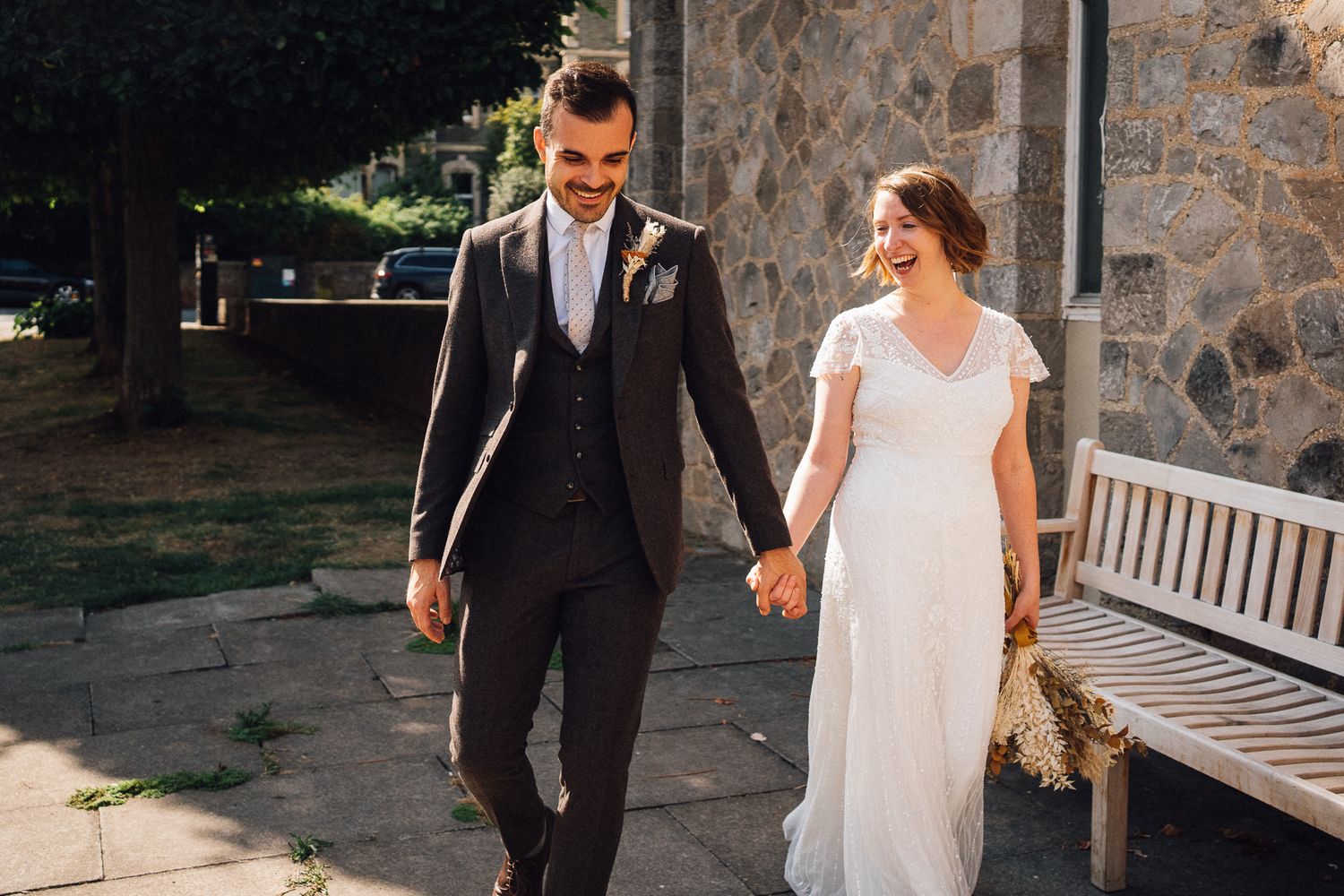 A couple walks hand in hand on their wedding day wearing a black suit and white dress near a stone wall and bench.