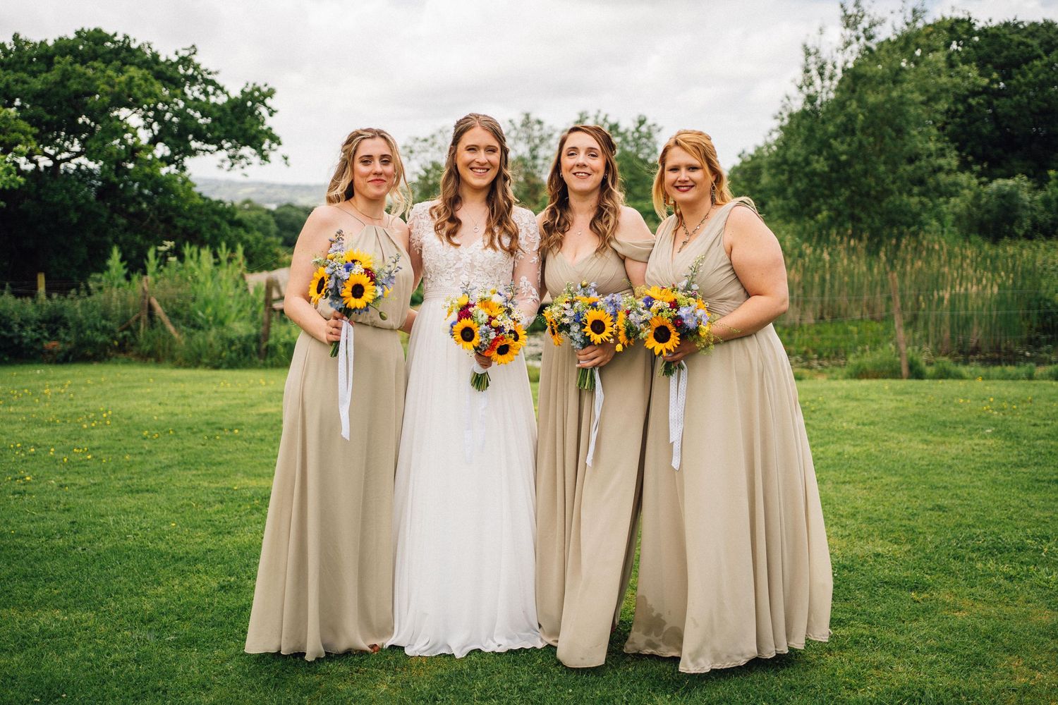 Group of women in formal dresses holding sunflower bouquets pose together on a green lawn.