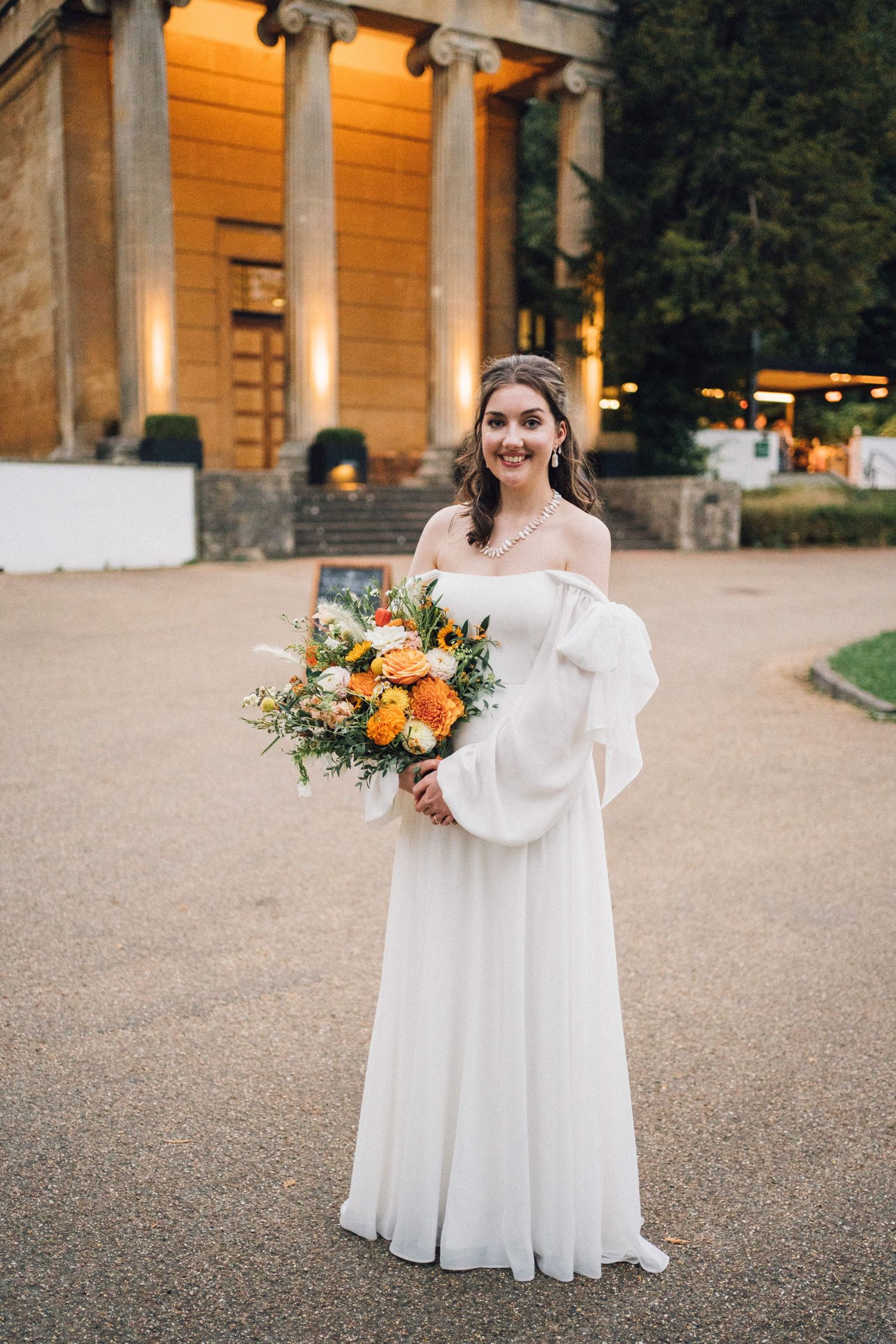 A bride in a flowing white dress poses outdoors against a warm-lit stone building backdrop.