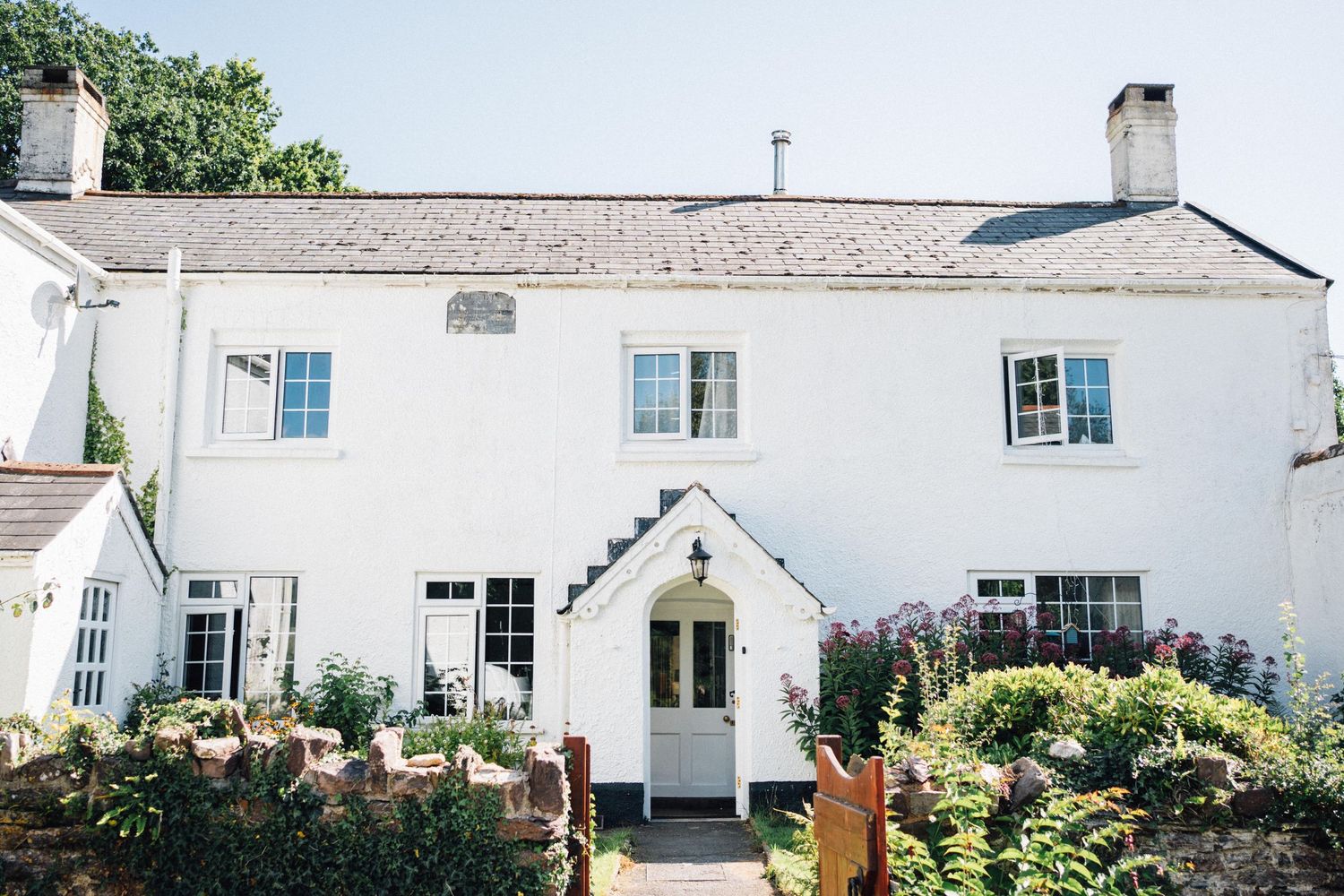 Charming white country cottage with blue shutters and a lush garden entrance featuring a welcoming covered porch.