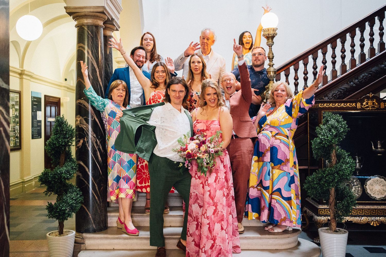 Wedding celebration group poses on elegant staircase with floral decorations and ornate railings.