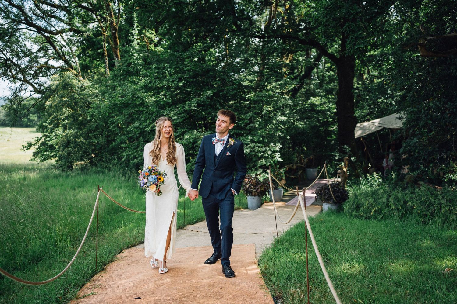 A couple walks along a garden path surrounded by lush greenery and natural landscaping during a wedding celebration.