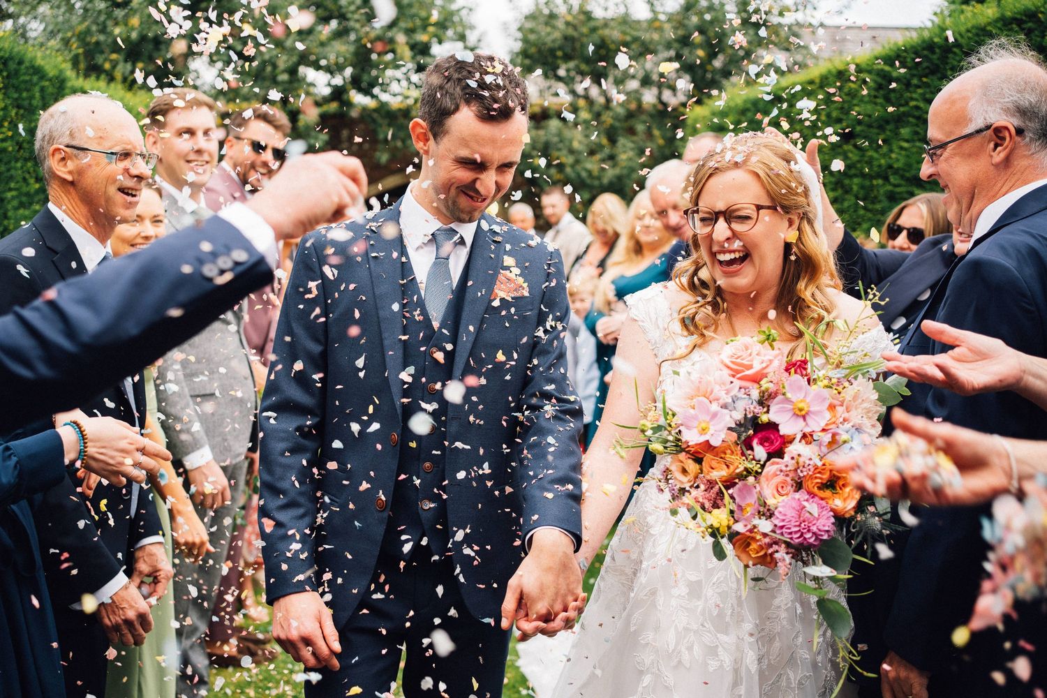 Wedding guests throw confetti over newlyweds in a garden setting with colorful summer flowers.