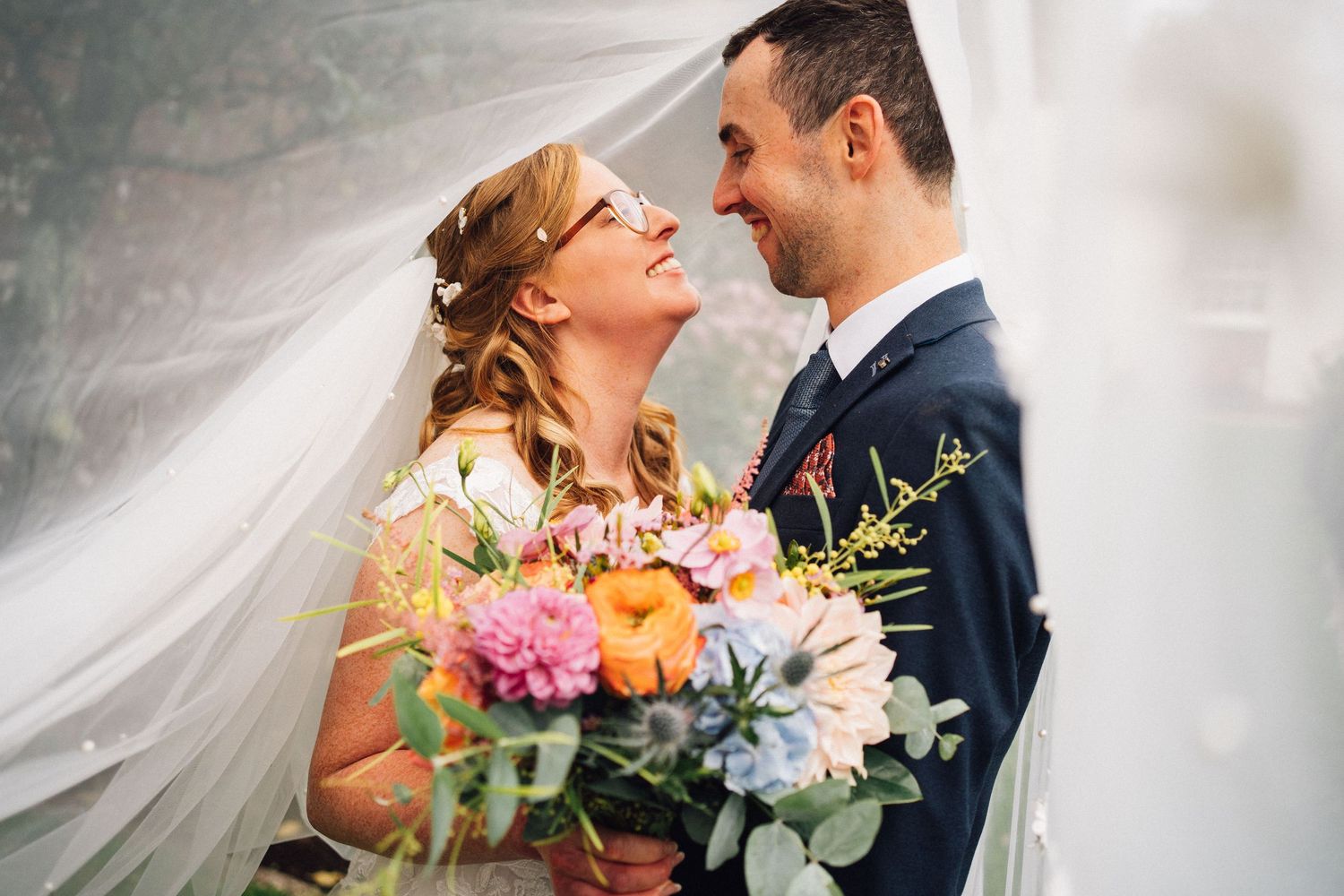 A romantic wedding couple shares an intimate moment under flowing white veils with colorful floral bouquets.