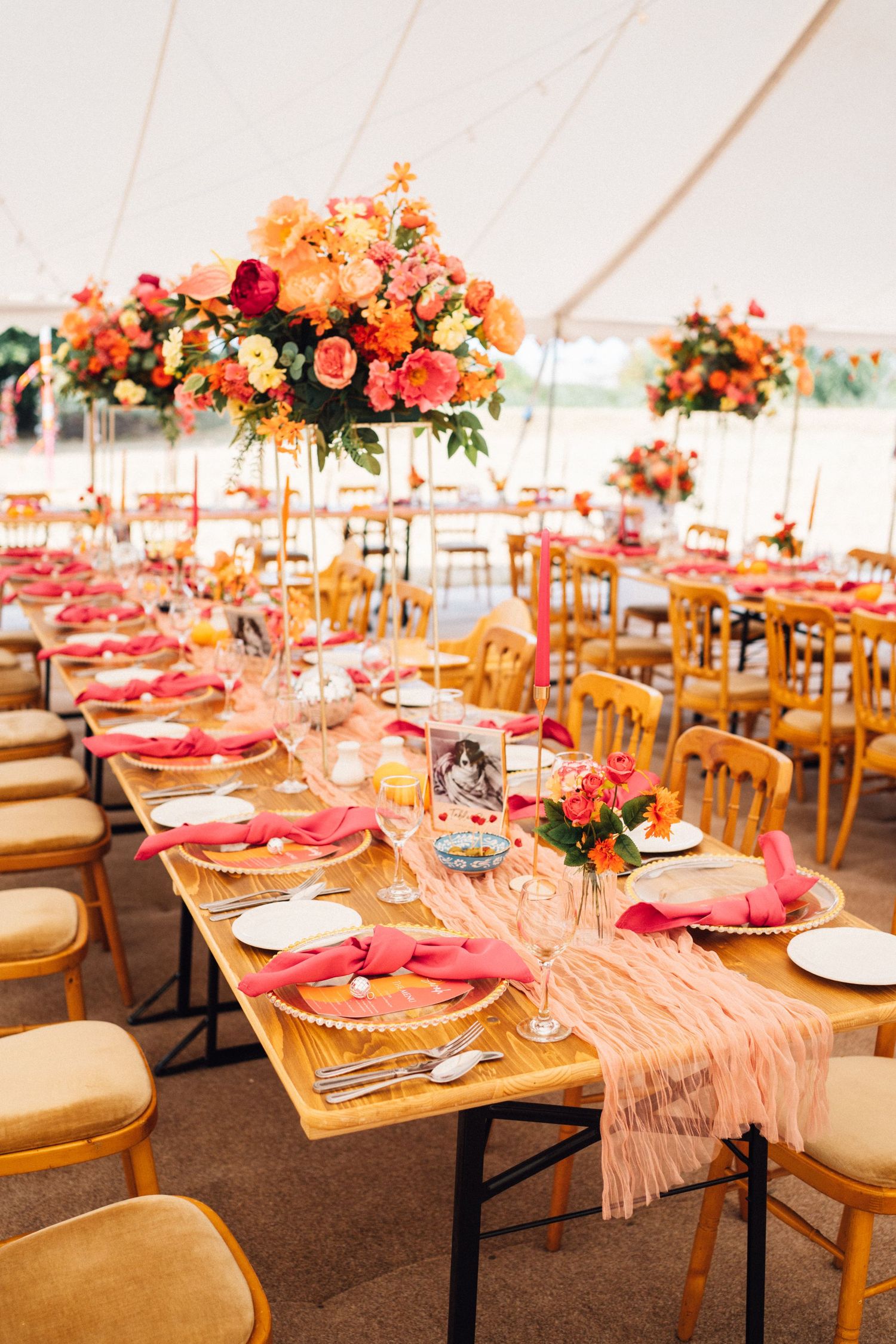 Elegant wedding reception table setting with coral napkins and floral centerpieces under a white tent.