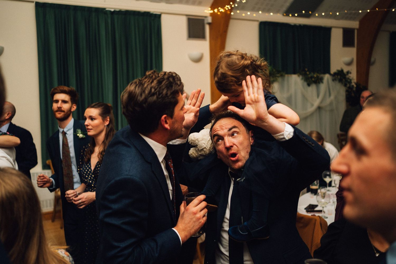Wedding reception party scene with guests dancing and celebrating under string lights and green curtains.