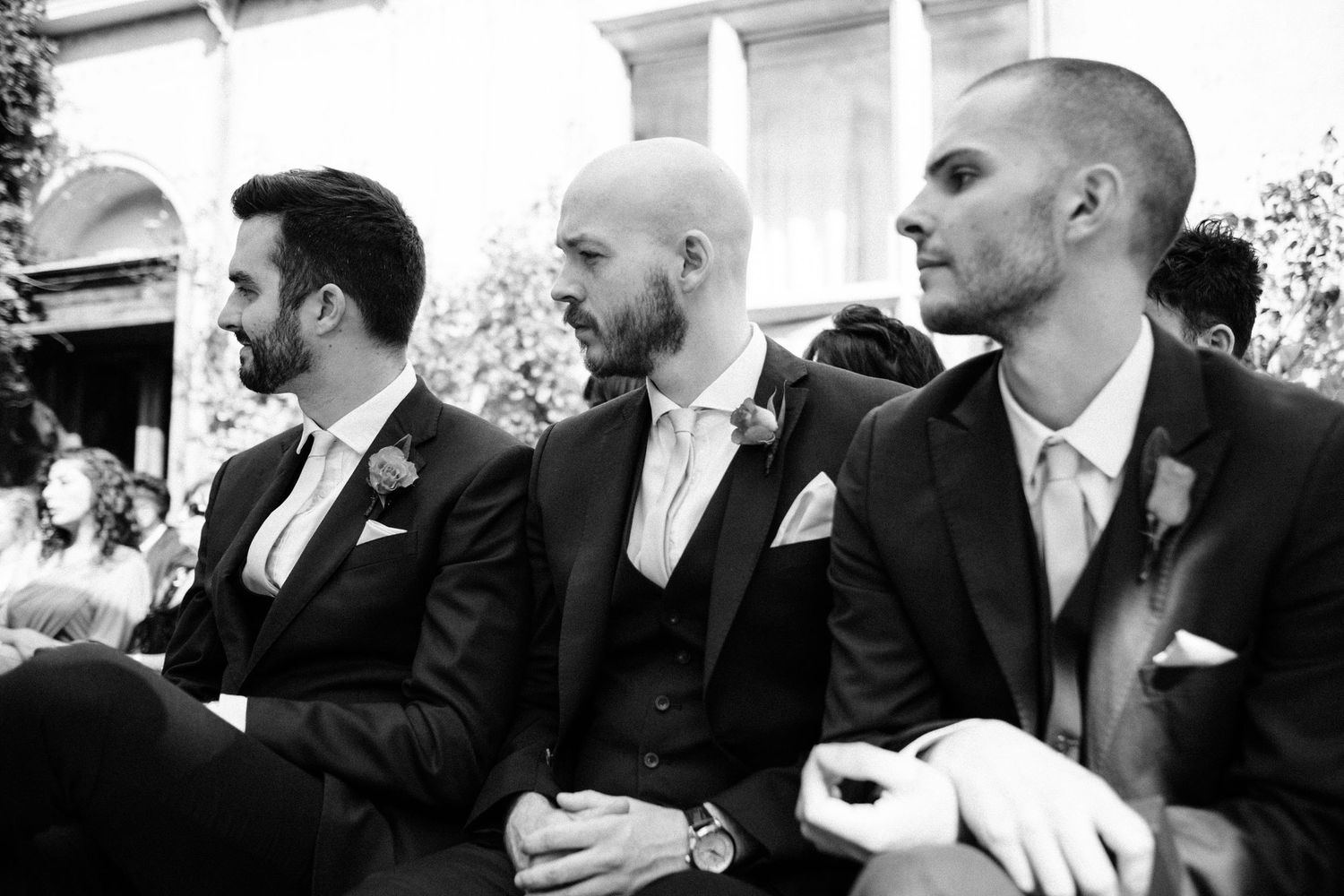 A black and white photo of groomsmen sitting in formal attire during a wedding ceremony.