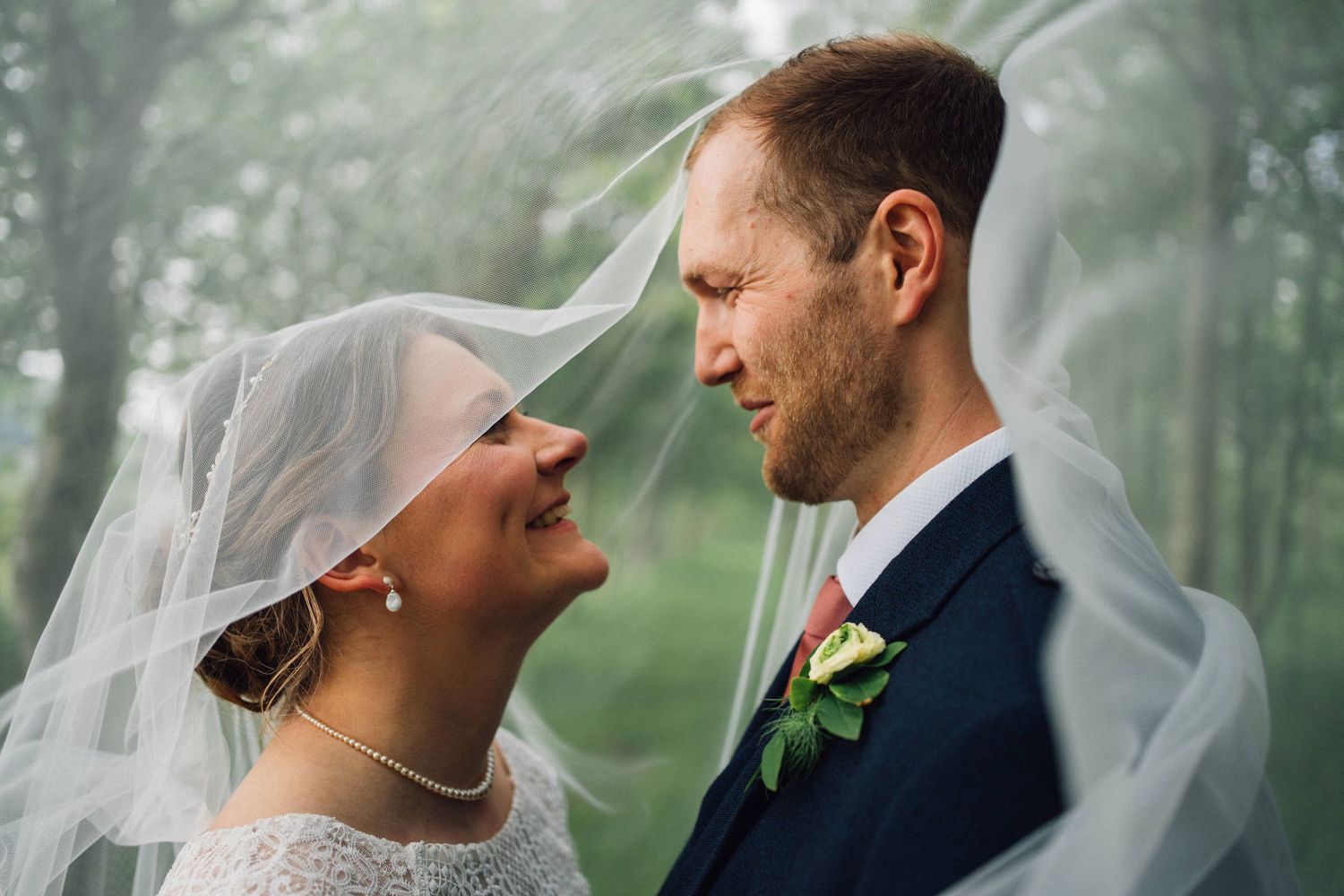 Intimate wedding portrait captured through flowing veil in misty forest setting.