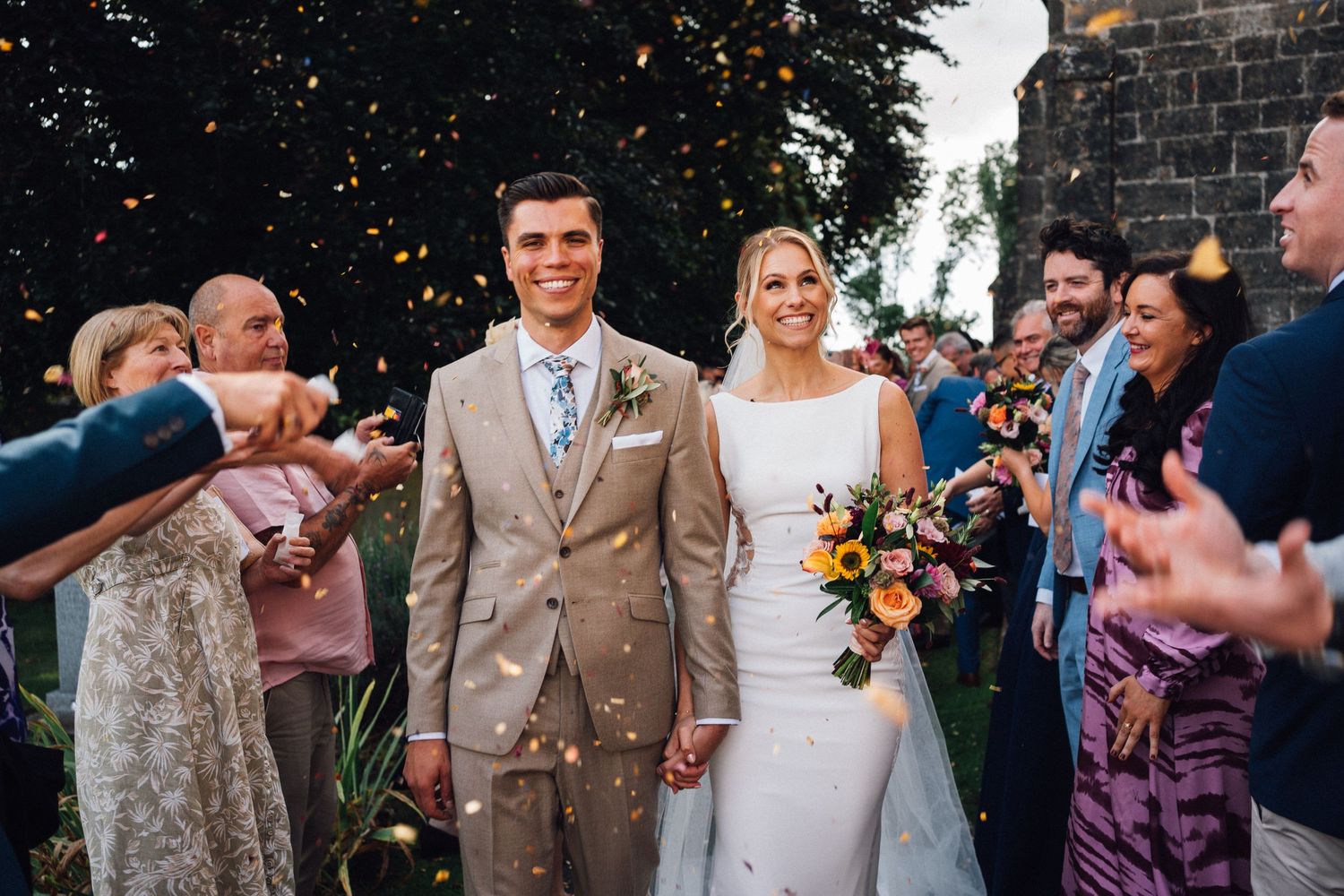 Wedding party celebrates outside historic stone church on sunny day.