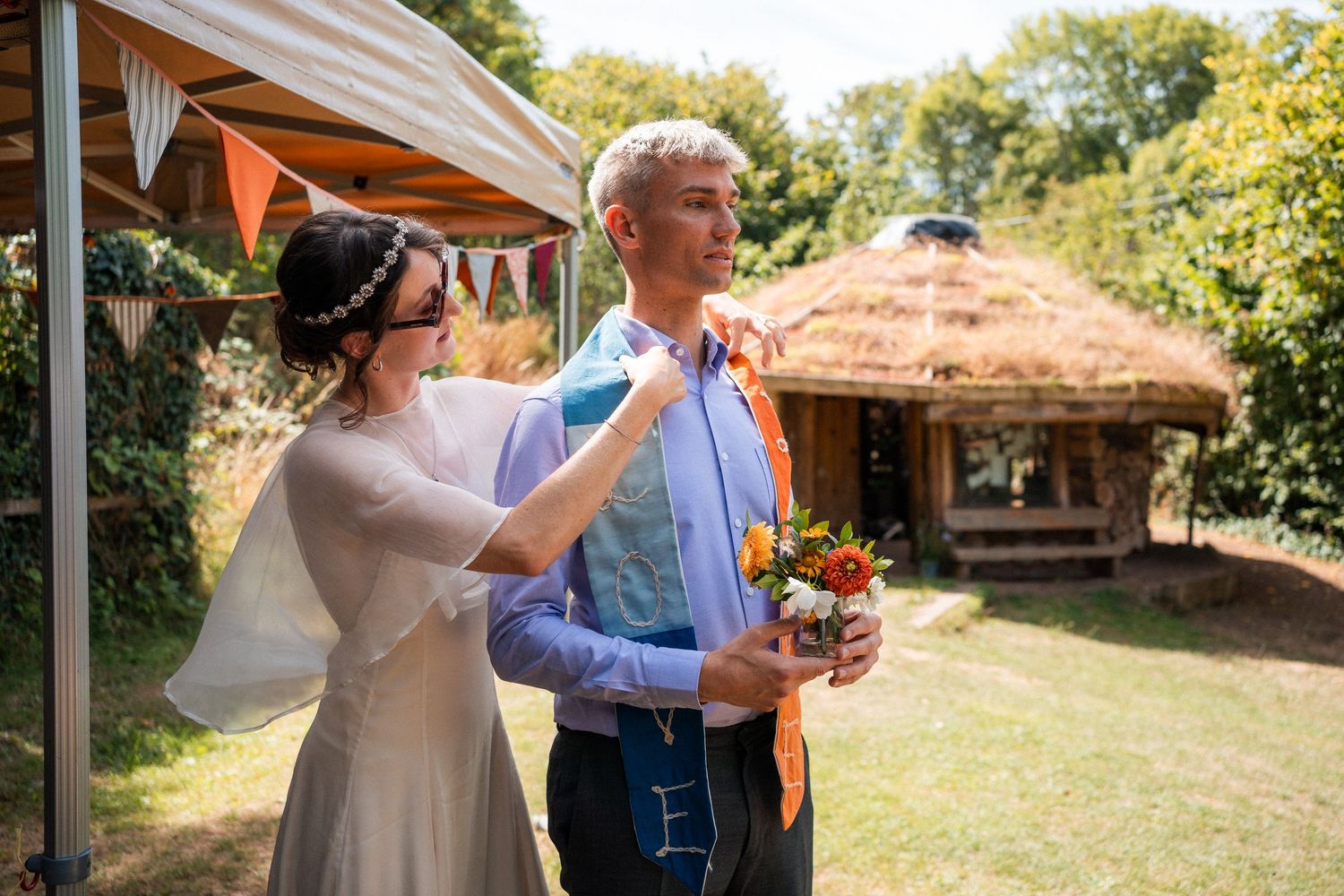 Bride adjusting groom's orange tie outdoors at rustic wedding venue with thatched roof building in background.