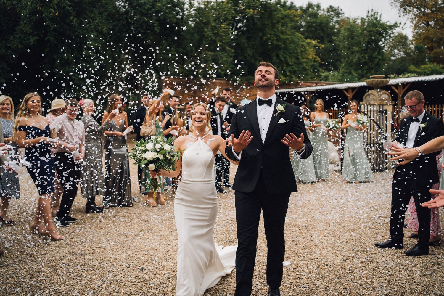 Wedding party walking through confetti on gravel path outside historic brick building.