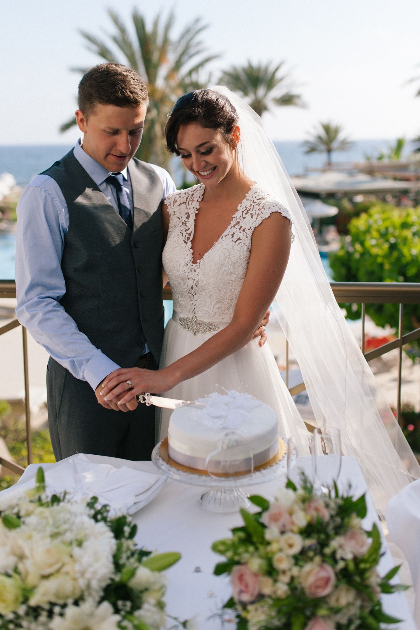 A couple cuts their wedding cake together outdoors with white floral decorations and palm trees in the background.