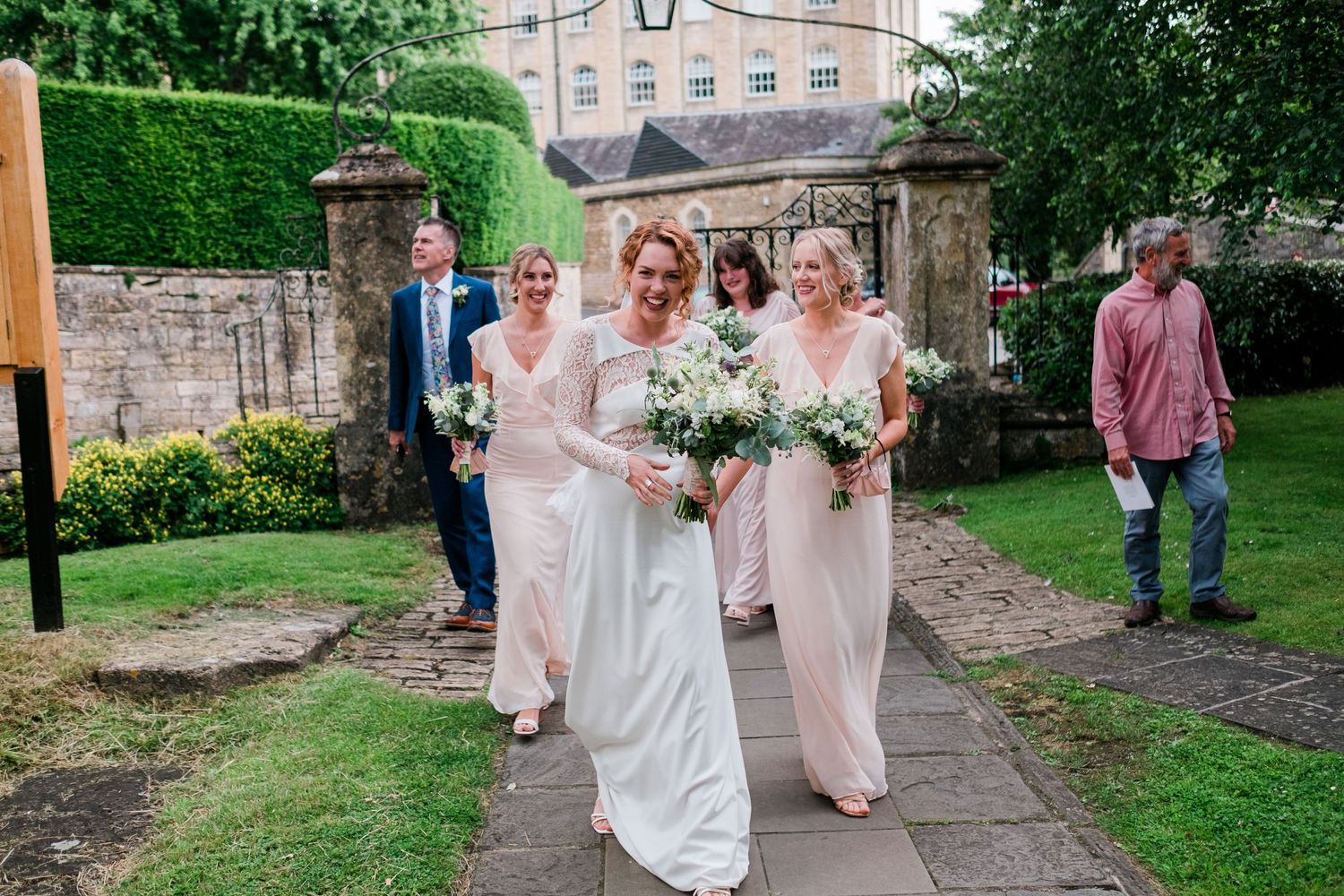 Wedding party walking along stone pathway in garden with historic building in background.