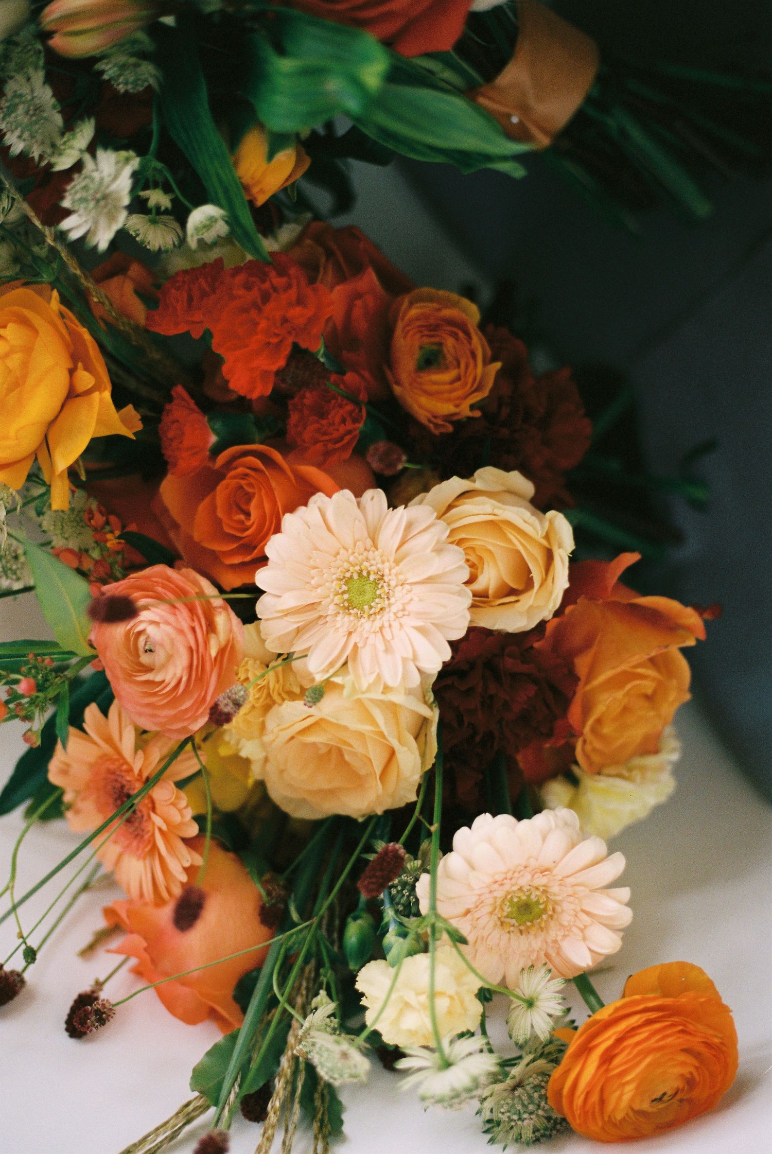 Lush autumn floral arrangement featuring orange roses, peach gerbera daisies, ranunculus, and deep red blooms with green foliage.