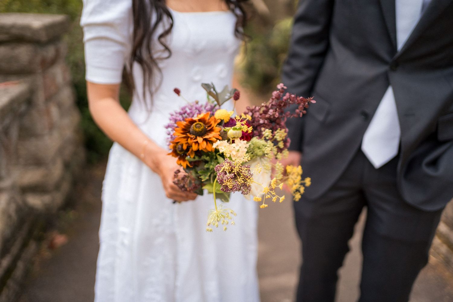 Bride in white dress and groom in gray suit holding a colorful wildflower wedding bouquet outdoors.