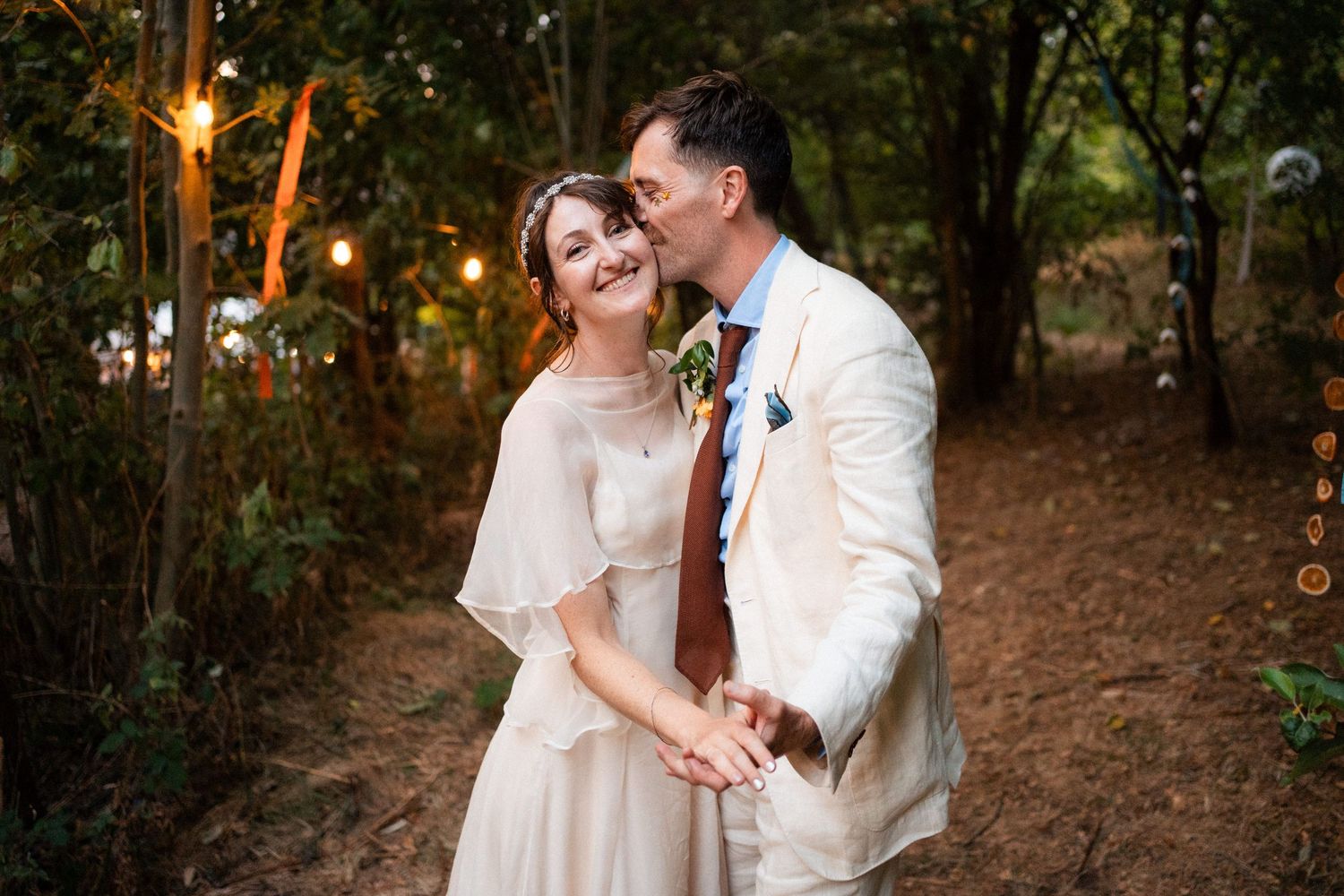 Bride and groom share a romantic moment in a woodland wedding setting with string lights glowing softly in the background.